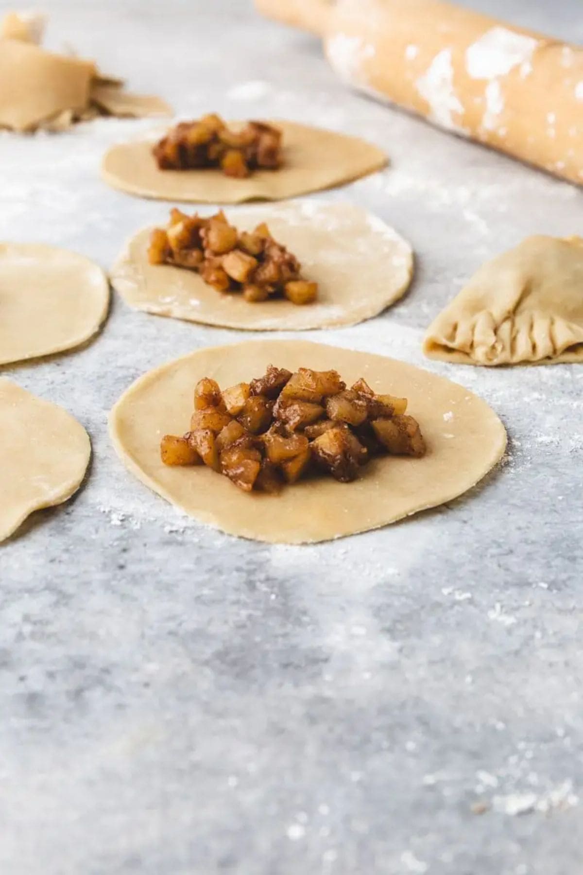 Unbaked dough circles topped with a diced fruit filling are on a floured surface, with a partially sealed pastry and a rolling pin in the background.