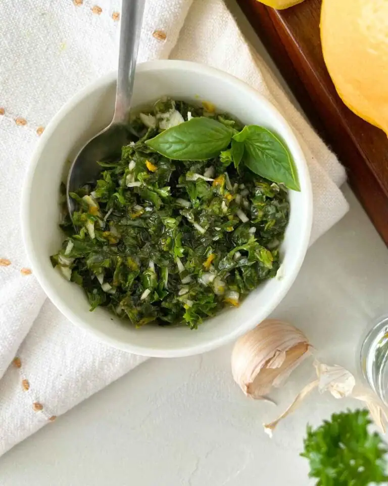 A small white bowl of finely chopped green herbs topped with basil leaves, with a metal spoon inside. Nearby are a garlic clove, parsley, a lemon, and a white cloth.