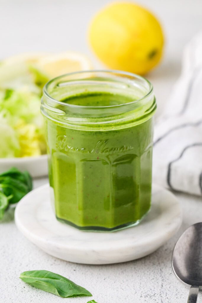 A glass jar filled with green salad dressing sits on a white coaster, making an inviting addition to any Food Photography Portfolio. In the background, a lemon, lettuce, and striped cloth create a fresh scene, while a basil leaf and spoon are artfully placed nearby.
