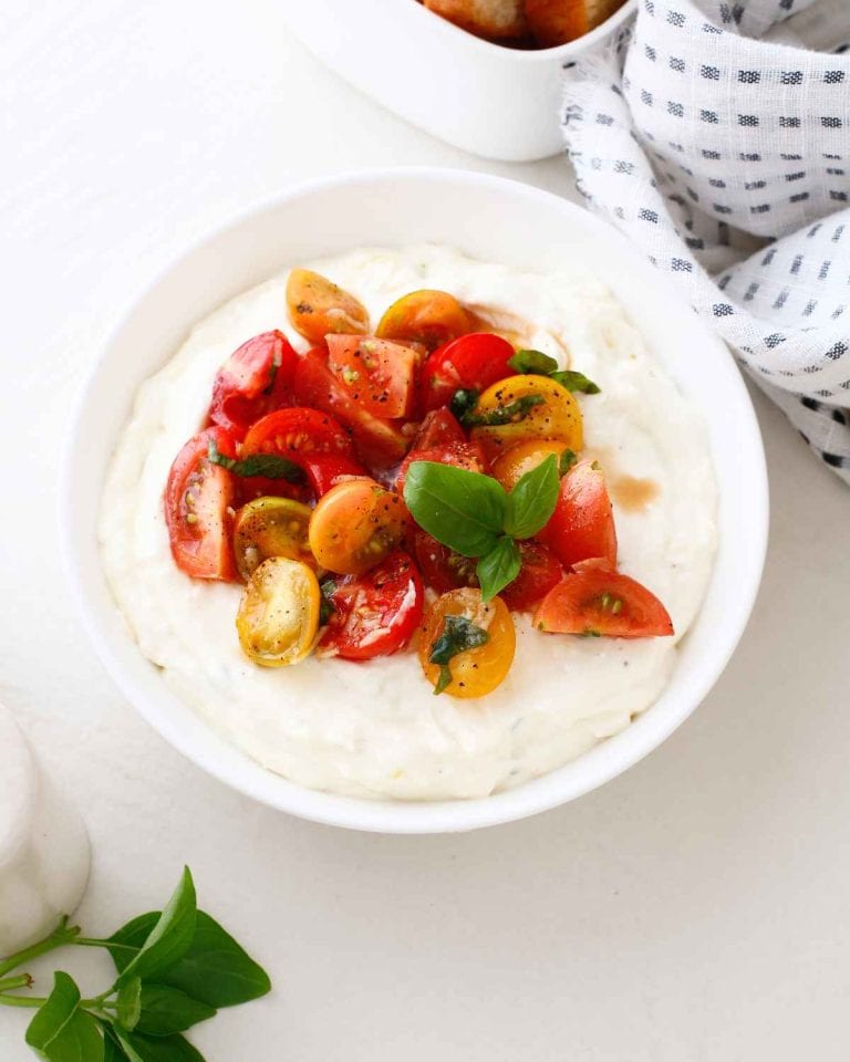 A white bowl filled with creamy dip, topped with chopped red and yellow cherry tomatoes, fresh basil, and black pepper. A checkered napkin and herbs are nearby on the white surface.