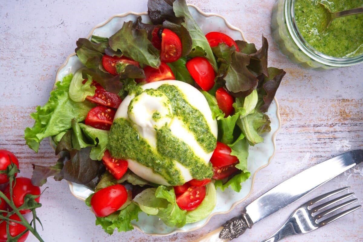 A plate with leafy greens, cherry tomatoes, and a ball of burrata cheese topped with green pesto. A jar of pesto, a fork, and a knife are nearby on a light surface.