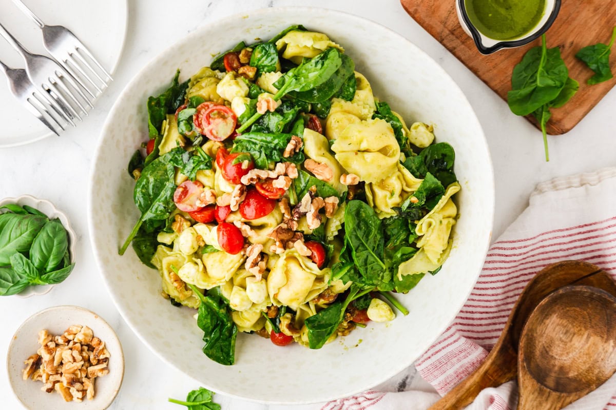 A large white bowl filled with tortellini pasta, fresh spinach, cherry tomatoes, and walnuts, sitting on a table with a small bowl of walnuts, wooden salad tongs, and a green dressing nearby.