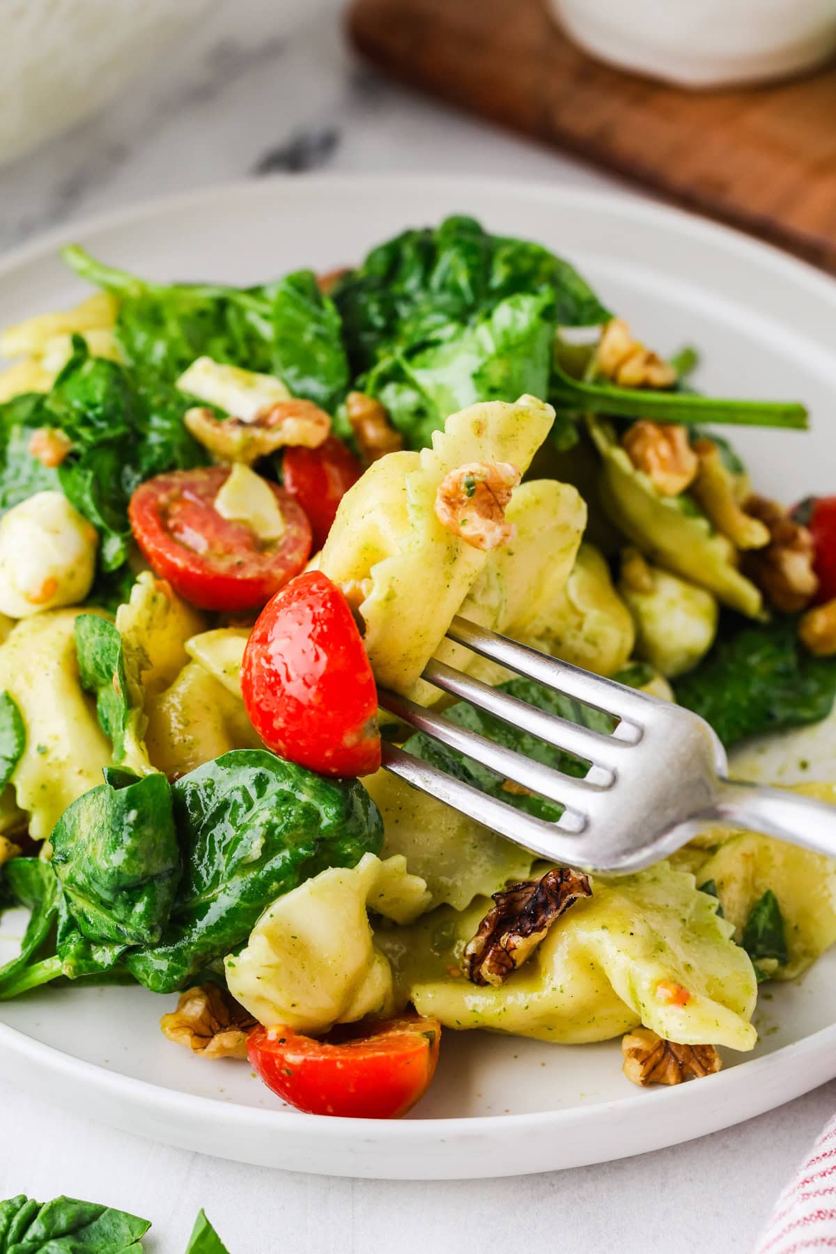 A fork holds a bite of tortellini pasta with fresh spinach, cherry tomatoes, walnuts, and chunks of cheese, all tossed in a light green pesto sauce and served on a white plate.