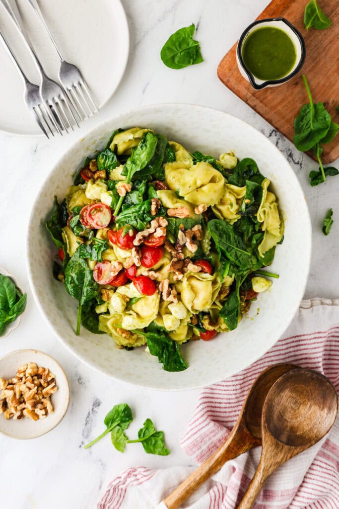 A bowl of tortellini salad with spinach, cherry tomatoes, and walnuts sits on a marble surface. Surrounding it are a small bowl of walnuts, wooden salad servers, a sauce jug, plates, and a striped napkin.