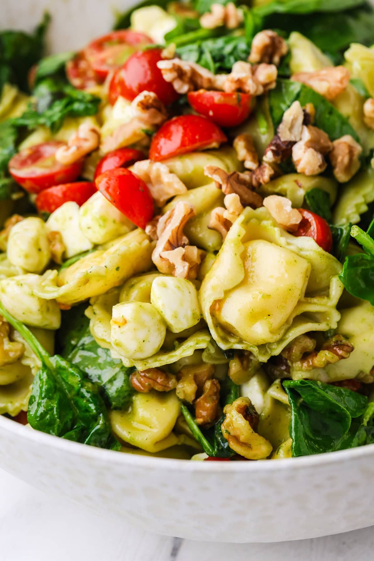 A close-up of a fresh salad with spinach, cheese tortellini, cherry tomatoes, mozzarella balls, walnuts, and pesto in a white bowl.