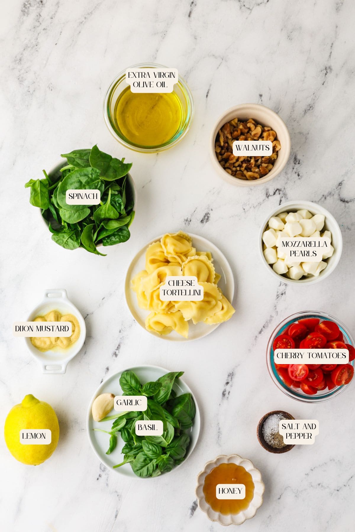 Flat lay of Caprese Tortellini Pasta Salad ingredients on marble: spinach, tortellini, mozzarella pearls, cherry tomatoes, walnuts, olive oil, lemon, basil, garlic, Dijon mustard, honey, and a bowl of salt and pepper.