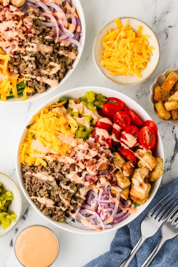 A vibrant showcase for any Food Photography Portfolio, this image features two bowls of salad topped with ground beef, shredded cheddar, sliced red onion, tomatoes, pickles, croutons, bacon bits, and creamy dressing. Sides of shredded cheese, croutons, pickles, and extra dressing are arranged nearby.