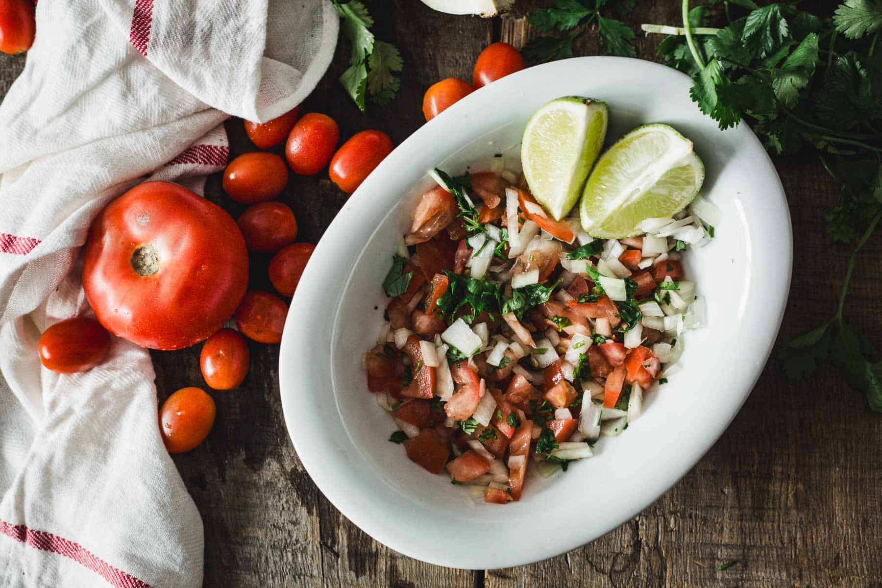 A white oval bowl filled with chopped tomatoes, onions, cilantro, and lime wedges sits on a rustic wooden table, surrounded by whole tomatoes, fresh cilantro, and a white towel with a red stripe.