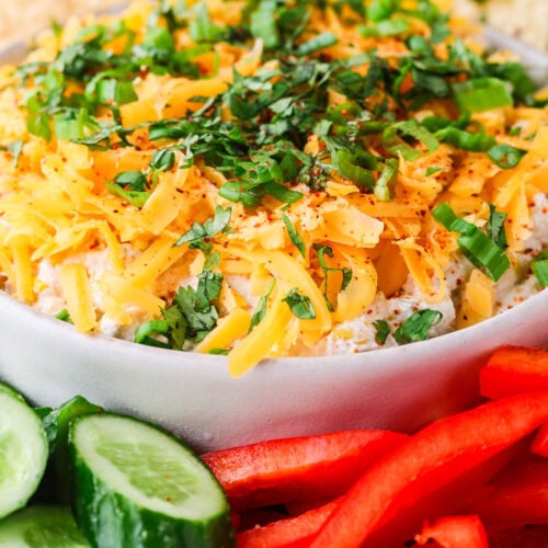 A bowl of creamy dip topped with shredded cheese and chopped herbs, surrounded by sliced cucumbers, red bell peppers, and tortilla chips.