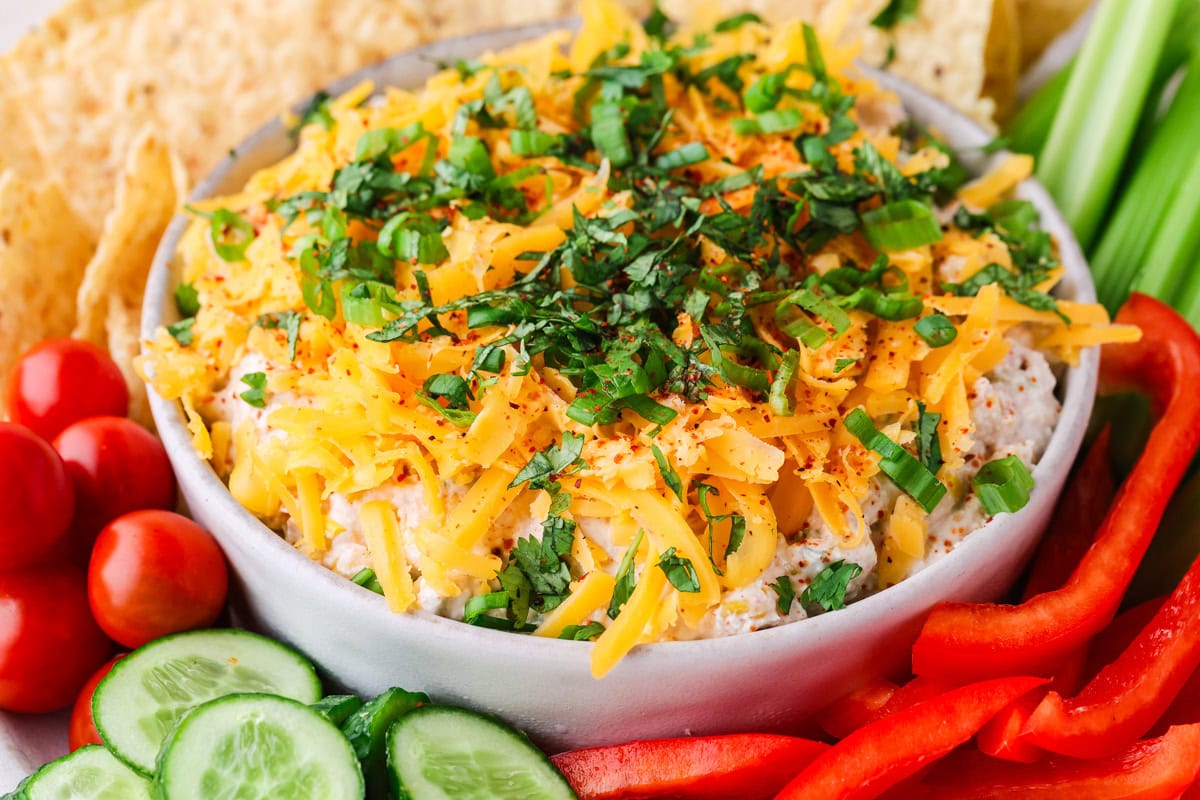 A bowl of dip topped with shredded cheese and herbs, surrounded by tortilla chips, celery sticks, cherry tomatoes, cucumber slices, and red bell pepper strips.