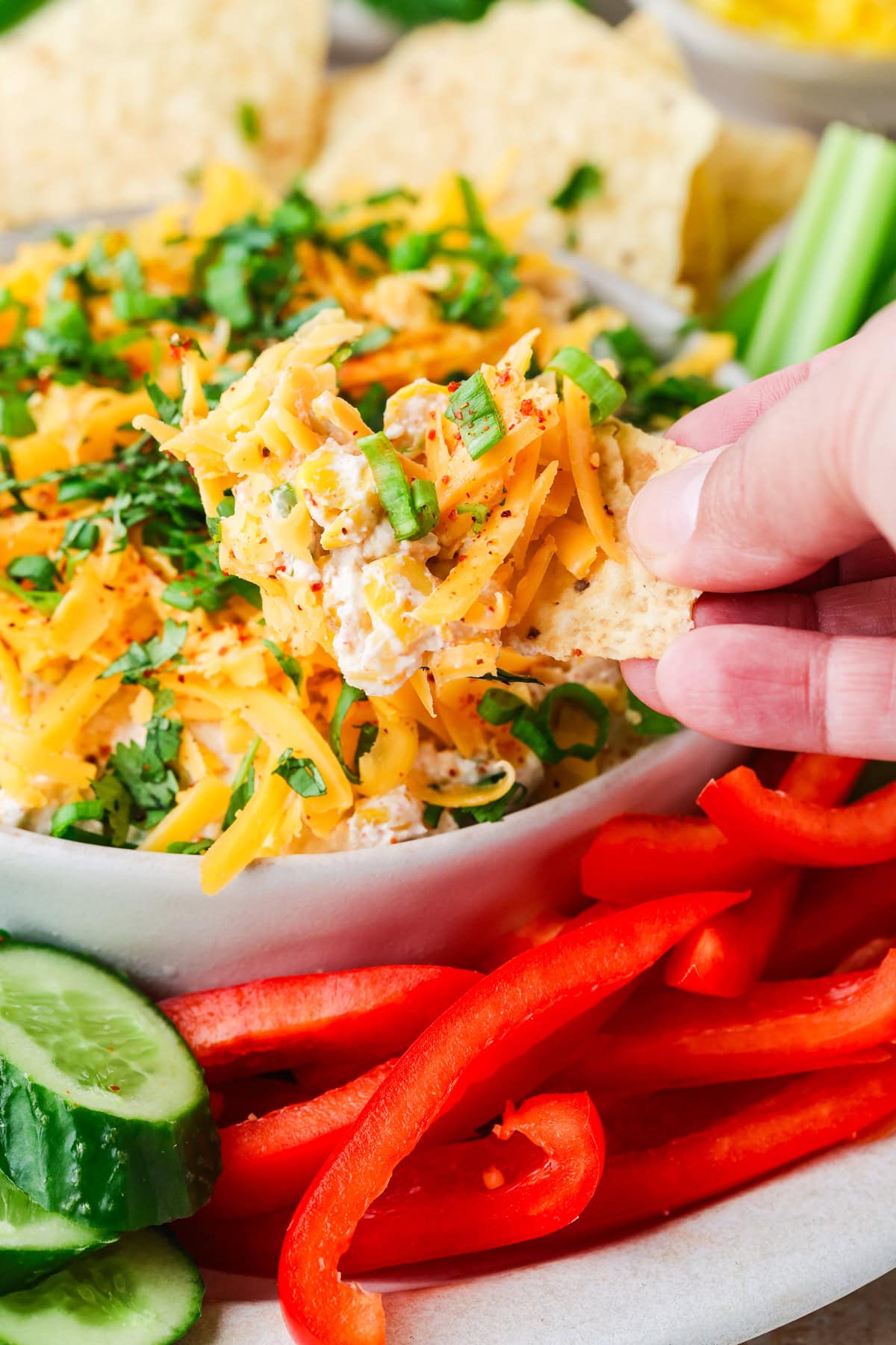 A hand dips a tortilla chip into a bowl of cheesy dip topped with shredded cheddar, herbs, and green onions. Sliced cucumbers and red bell peppers are on the side.