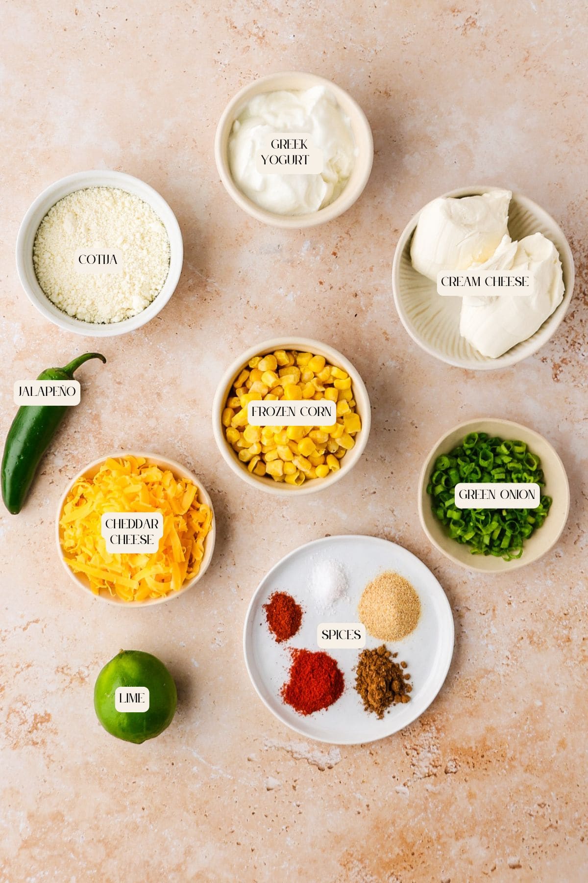 Seven bowls with labeled ingredients: cotija cheese, Greek yogurt, cream cheese, cheddar cheese, frozen corn, green onion, plus whole jalapeño and lime, and a plate with assorted spices on a light countertop.