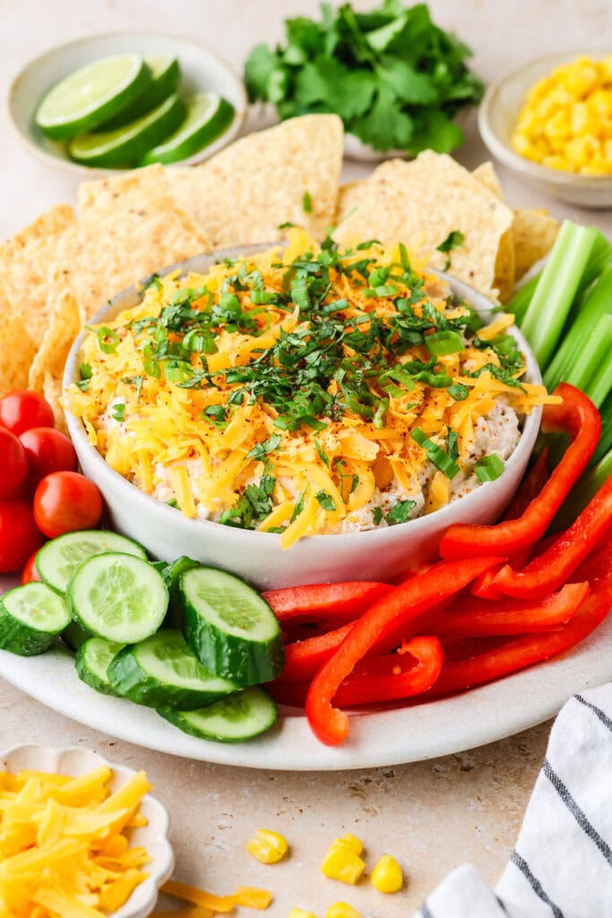 A bowl of dip topped with shredded cheese and herbs, surrounded by tortilla chips, cucumber slices, cherry tomatoes, red bell pepper strips, celery sticks, and lime wedges. Corn and cilantro are in small bowls nearby.