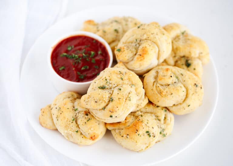 A plate of golden-brown garlic knots garnished with herbs, served alongside a small bowl of marinara sauce topped with chopped parsley.