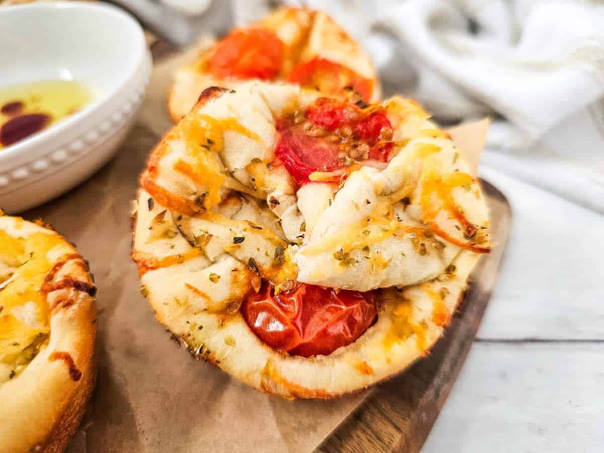 A close-up of a mini savory tart with melted cheese, tomatoes, and herbs on a wooden board, next to a bowl of olive oil for dipping.