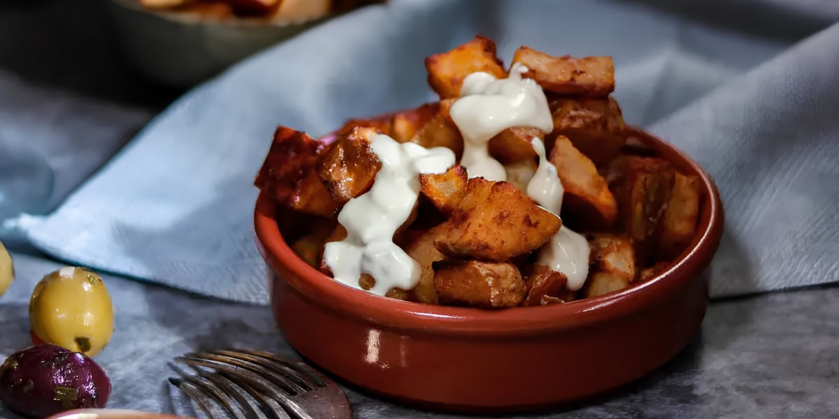 A brown dish filled with golden, crispy potato cubes topped with a white creamy sauce sits on a table next to a fork and some olives.