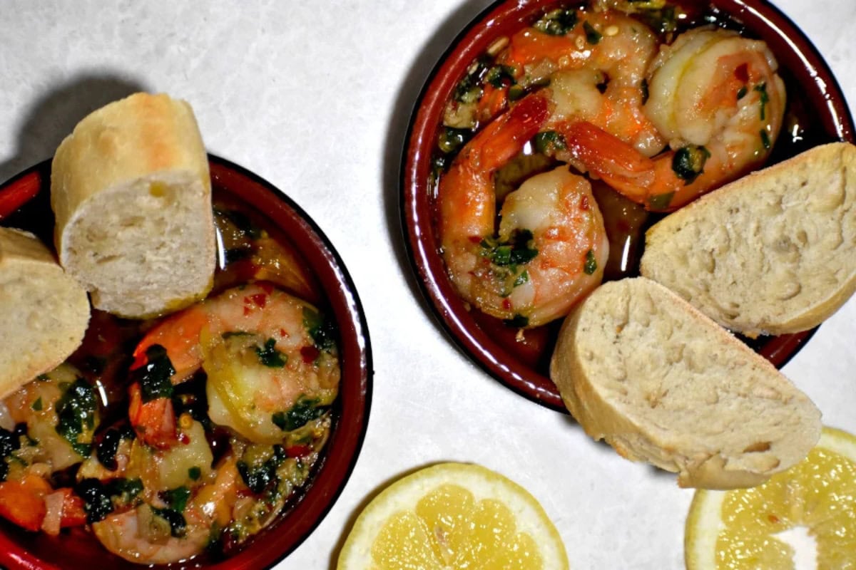 Two bowls of shrimp in a garlic herb sauce, each served with slices of crusty bread. Lemon slices are placed beside the bowls on a white surface.