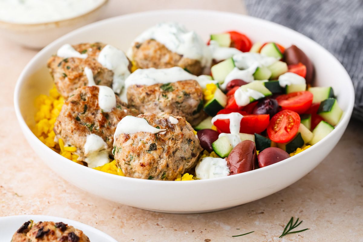 A bowl with yellow rice, Greek Chicken Meatballs topped with white sauce, cherry tomatoes, cucumber, olives, and zucchini, with a small dish of sauce and a gray napkin in the background.