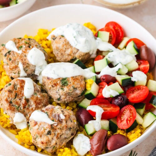 A bowl with yellow rice, five Greek Chicken Meatballs, sliced cucumbers, cherry tomatoes, Kalamata olives, and white sauce drizzled on top. A small bowl of white sauce is in the background.