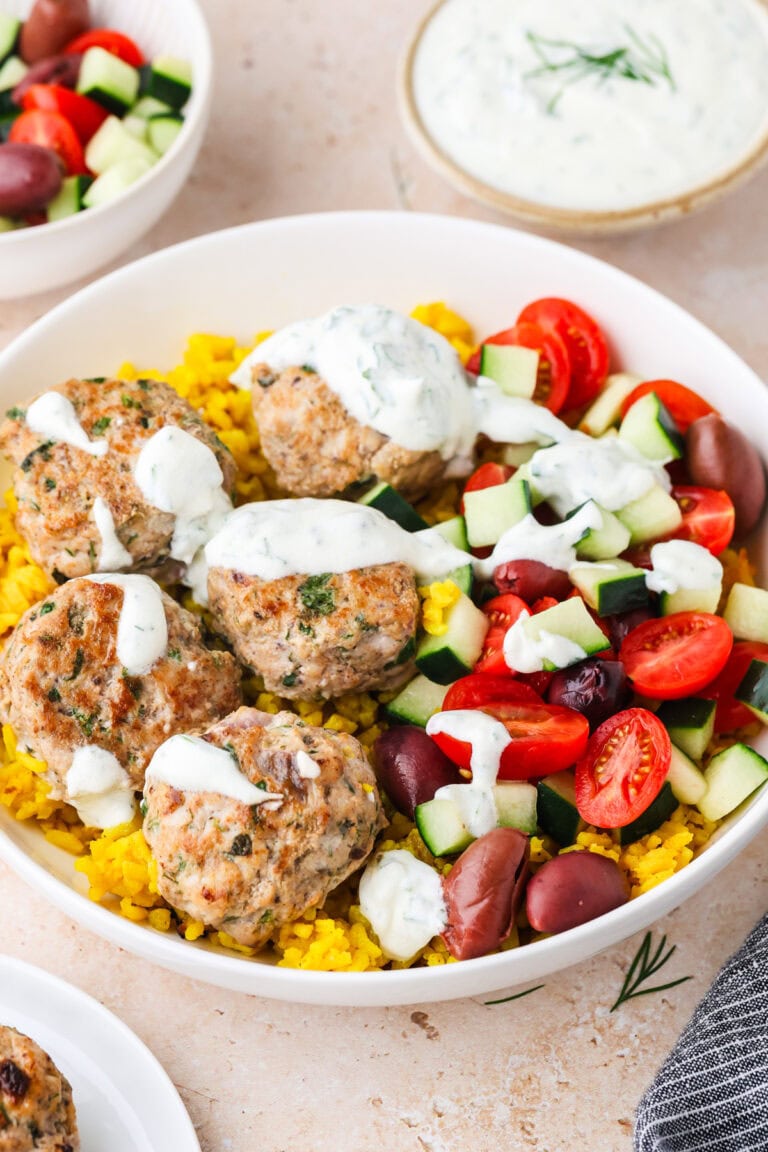 A bowl with yellow rice, five Greek Chicken Meatballs, sliced cucumbers, cherry tomatoes, Kalamata olives, and white sauce drizzled on top. A small bowl of white sauce is in the background.