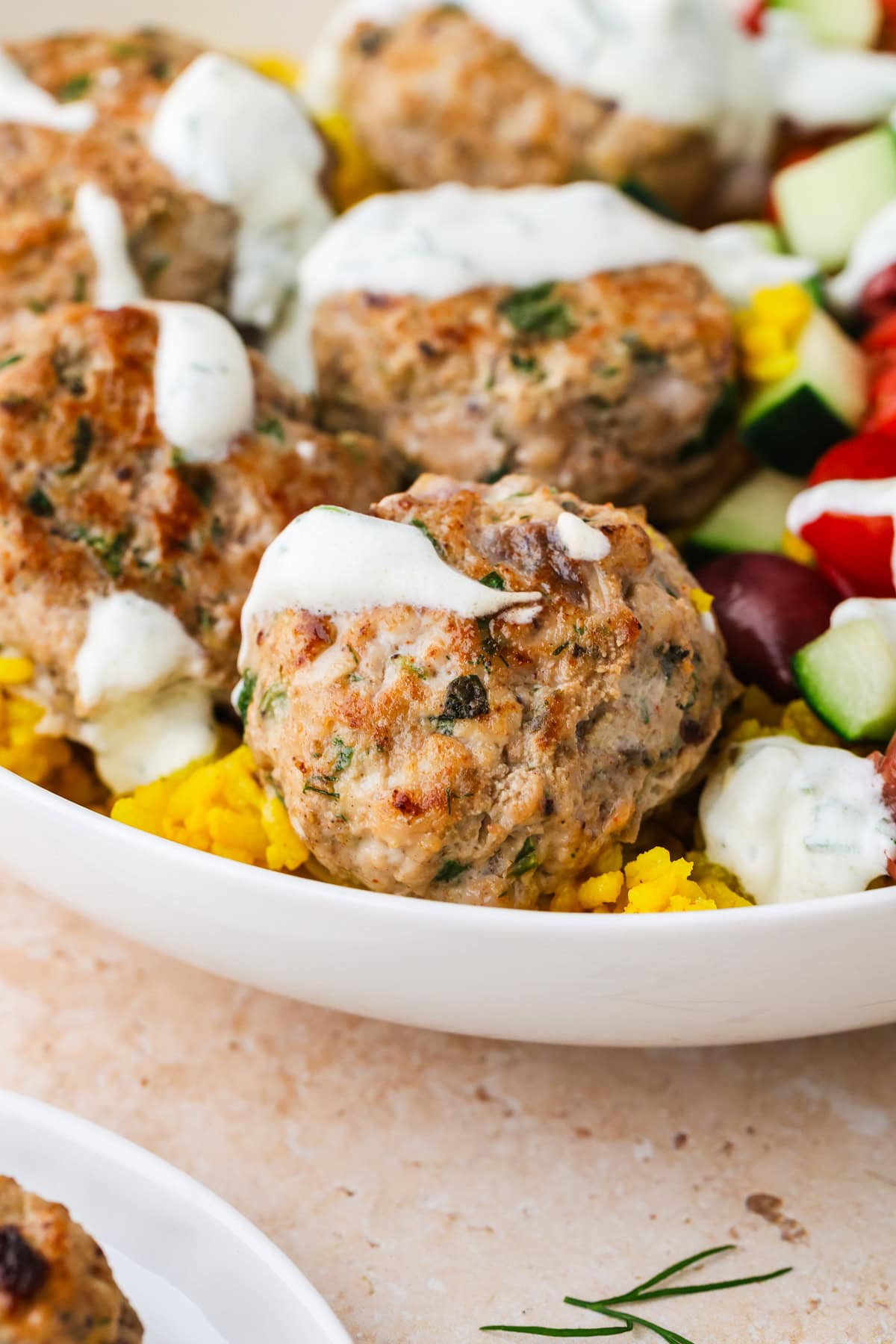 Greek Chicken Meatballs topped with white sauce rest on yellow rice, accompanied by chopped cucumber, tomato, and olives in a white bowl.