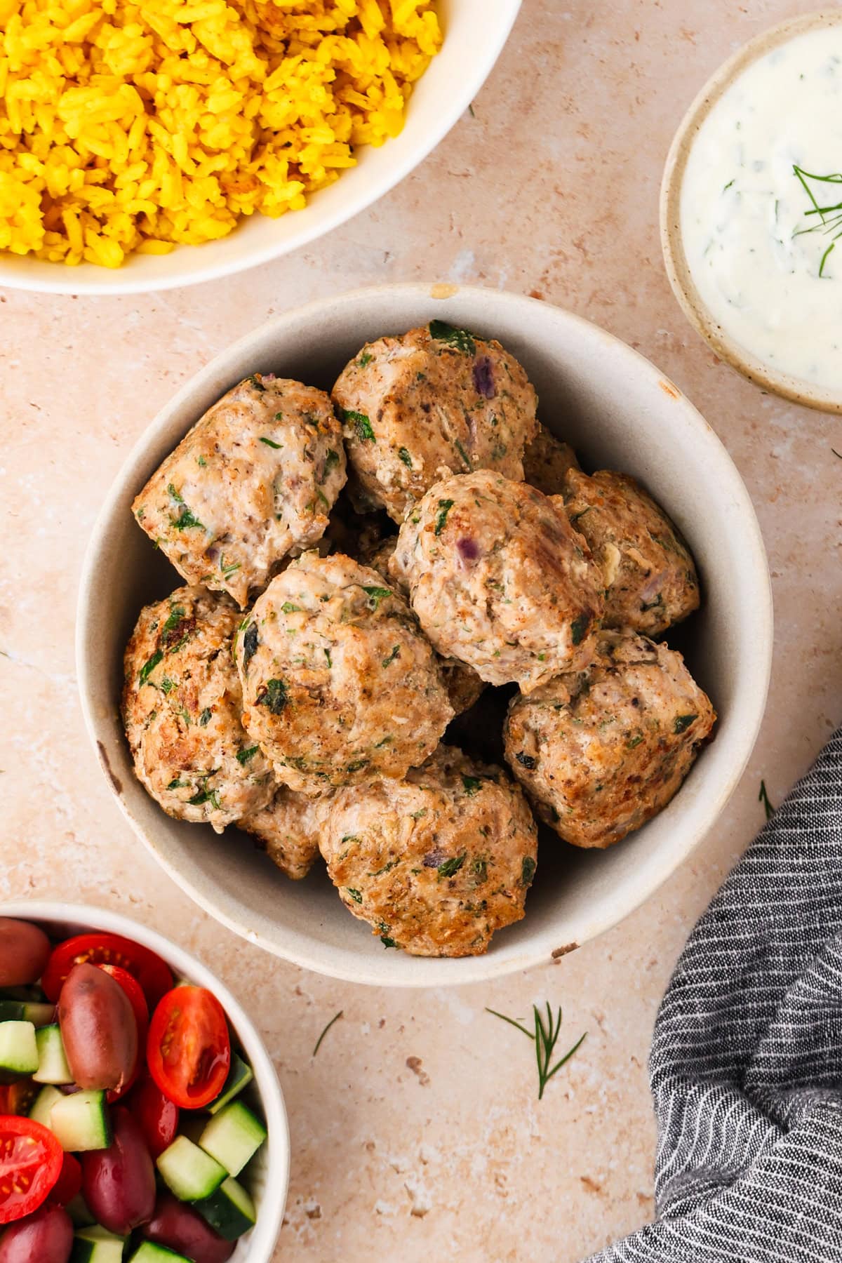 A bowl of Greek Chicken Meatballs sits on a table with a dish of yellow rice, a bowl of cucumber salad with tomatoes and olives, and a small bowl of creamy white sauce nearby.