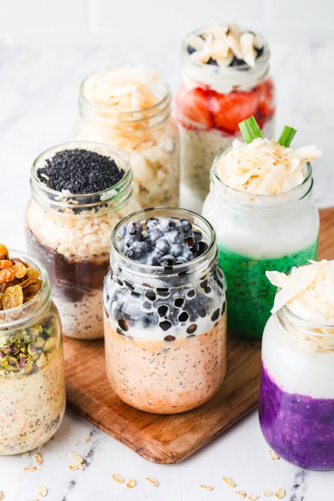 A vibrant Food Photography Portfolio image showcases seven jars filled with colorful overnight oats, each topped with an array of fruits, seeds, coconut flakes, and nuts, all beautifully arranged on a wooden board against a light background.