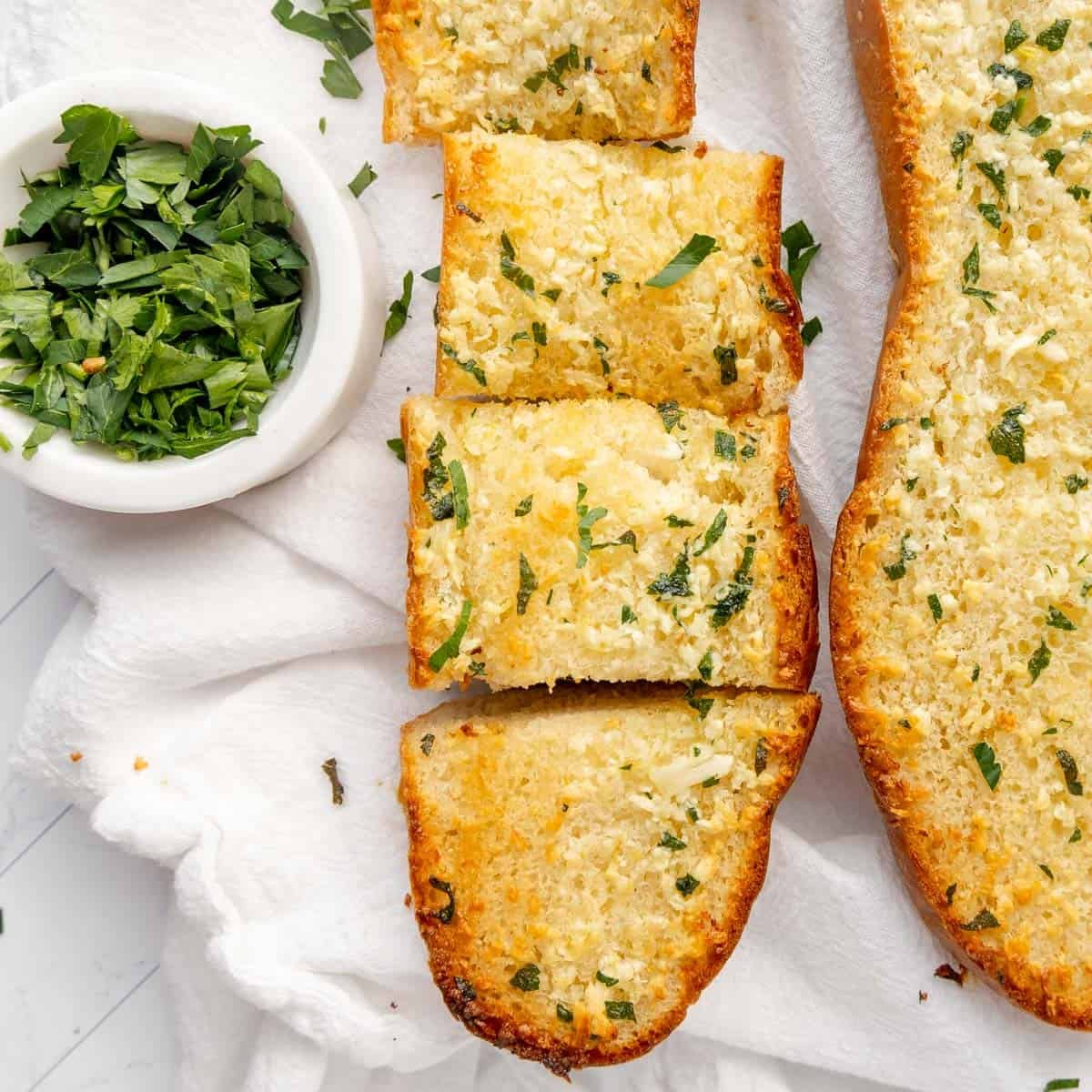 Sliced garlic bread topped with chopped parsley on a white cloth, with a small bowl of fresh green herbs beside it.