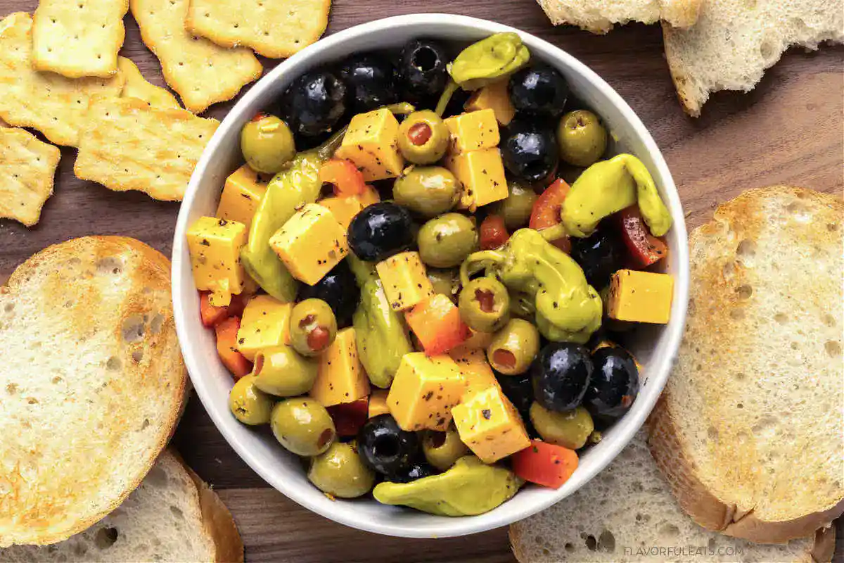 A bowl of antipasto salad with green and black olives, cheddar cheese cubes, pepperoncini, and red bell peppers, surrounded by slices of toasted bread and crackers.