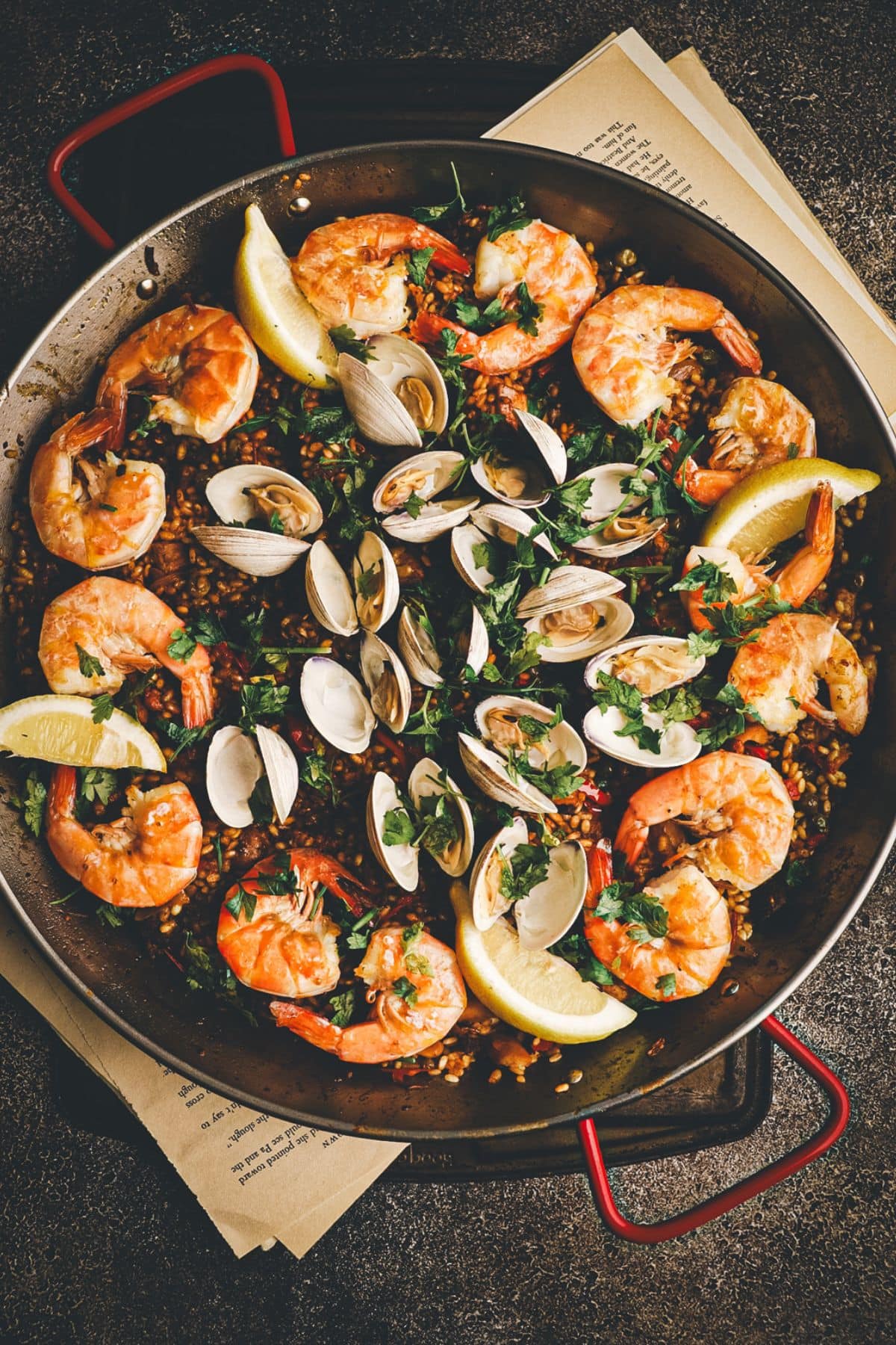 A large pan of seafood paella topped with shrimp, clams, lemon wedges, and fresh herbs, set on a dark surface with pieces of paper underneath for a paella side dishes round up.