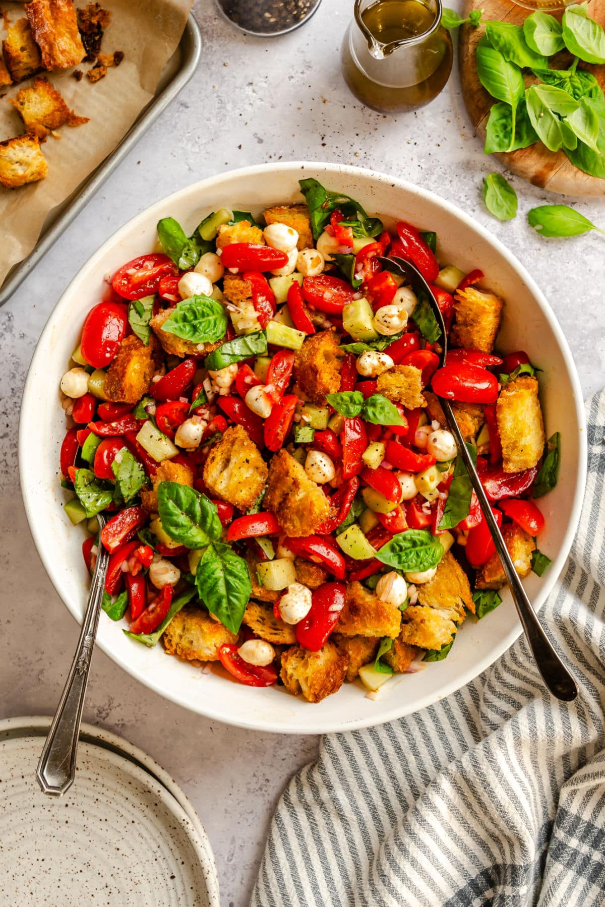 A bowl of panzanella salad with cherry tomatoes, mozzarella balls, basil, cucumbers, and toasted bread cubes. Two serving spoons are in the bowl, with olive oil, basil leaves, and a striped napkin nearby.