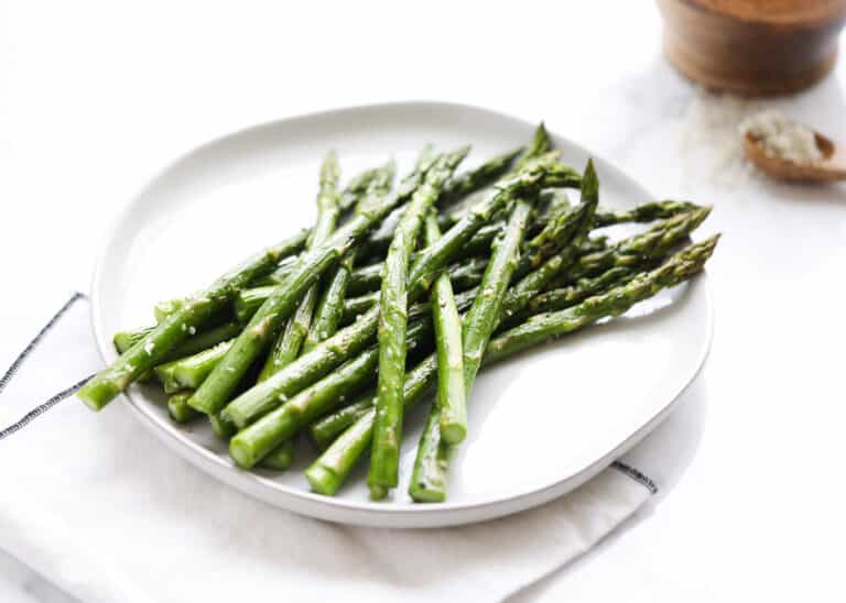 A white plate with cooked asparagus spears sprinkled with coarse salt, placed on a white napkin. A wooden bowl and spoon with salt are blurred in the background.