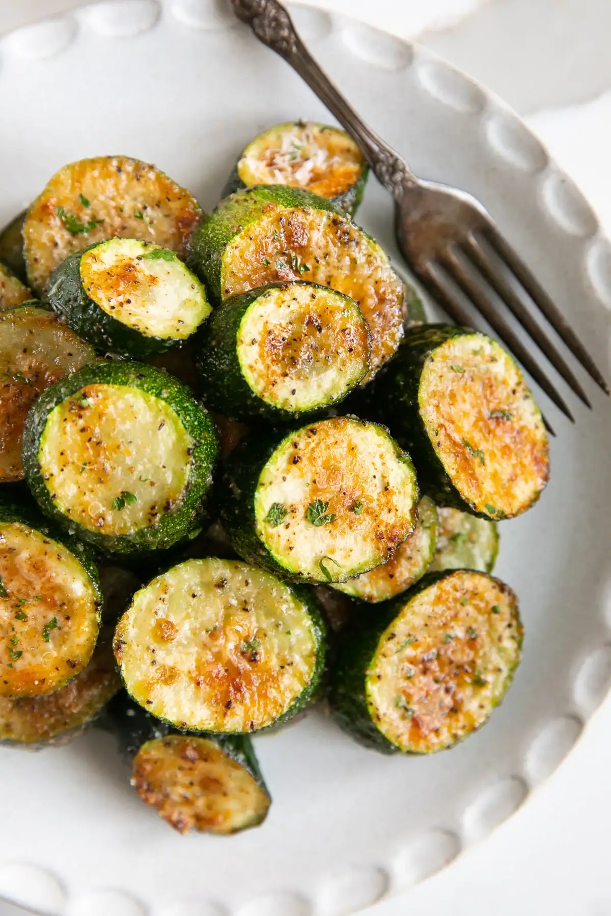 Sliced roasted zucchini rounds seasoned with herbs and spices, arranged on a white plate with a fork beside them.