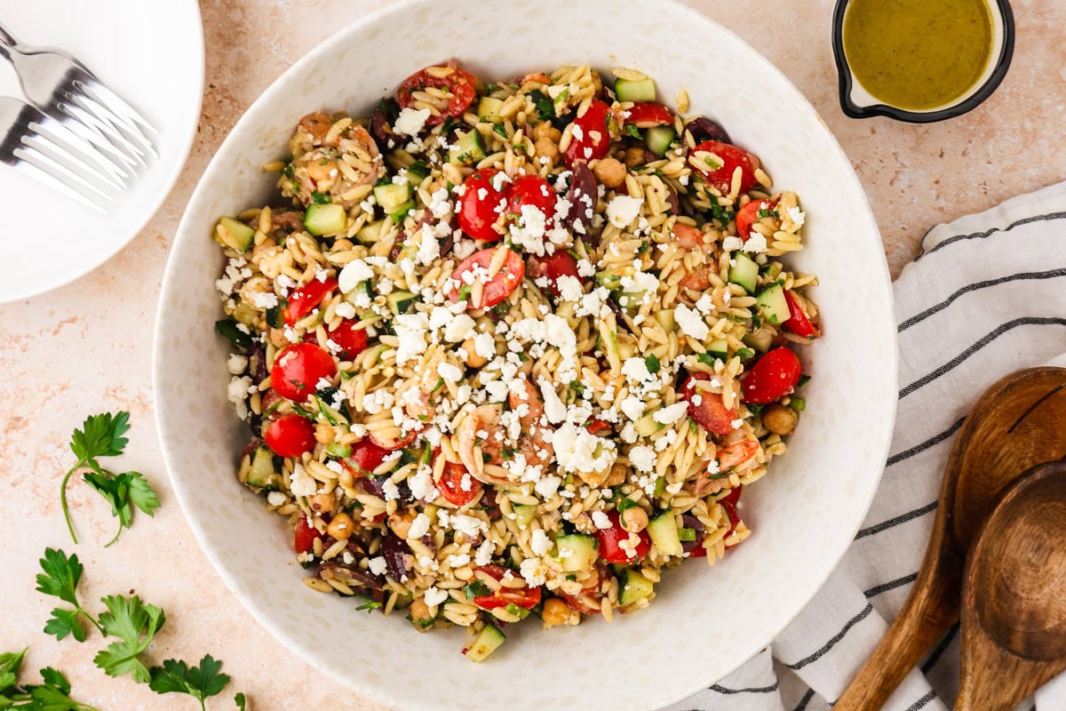 A large bowl of Shrimp Orzo Salad with cherry tomatoes, cucumbers, olives, chickpeas, and crumbled feta cheese sits next to a small bowl of dressing, plates, forks, and wooden serving utensils.