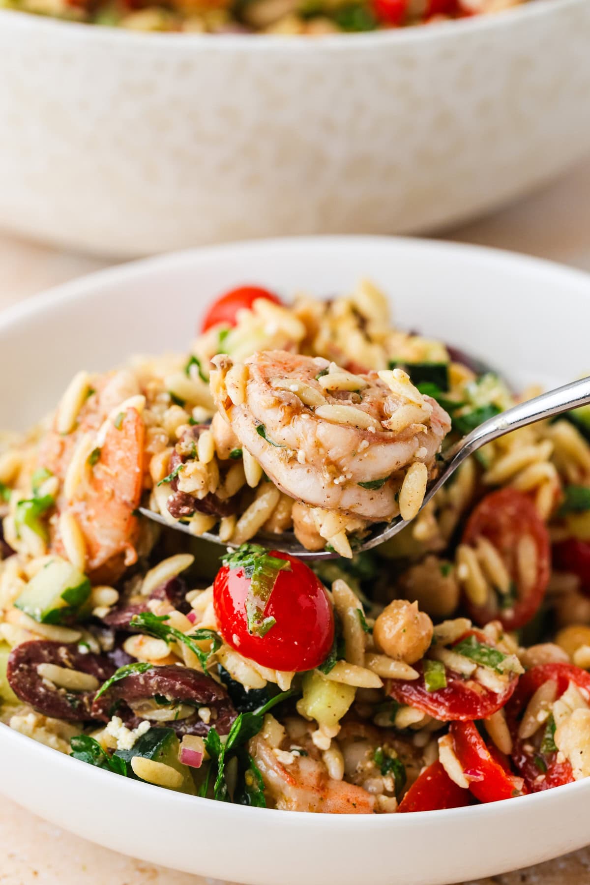 A close-up of a fork holding Shrimp Orzo Salad with tomatoes, cucumber, chickpeas, olives, spinach, and feta in a white bowl.