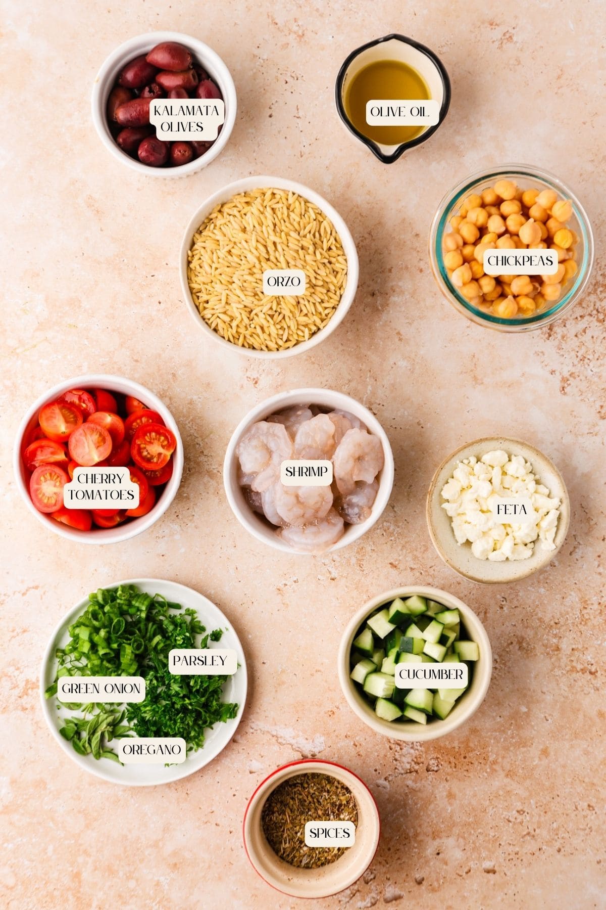 Top-down view of bowls containing all the fresh ingredients for a Shrimp Orzo Salad—kalamata olives, olive oil, chickpeas, cherry tomatoes, shrimp, feta, cucumber, green onion, parsley, oregano, and spices—all labeled on a light surface.