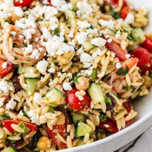 A close-up of a bowl filled with Shrimp Orzo Salad, featuring cherry tomatoes, cucumbers, olives, chickpeas, fresh herbs, and crumbled feta cheese.