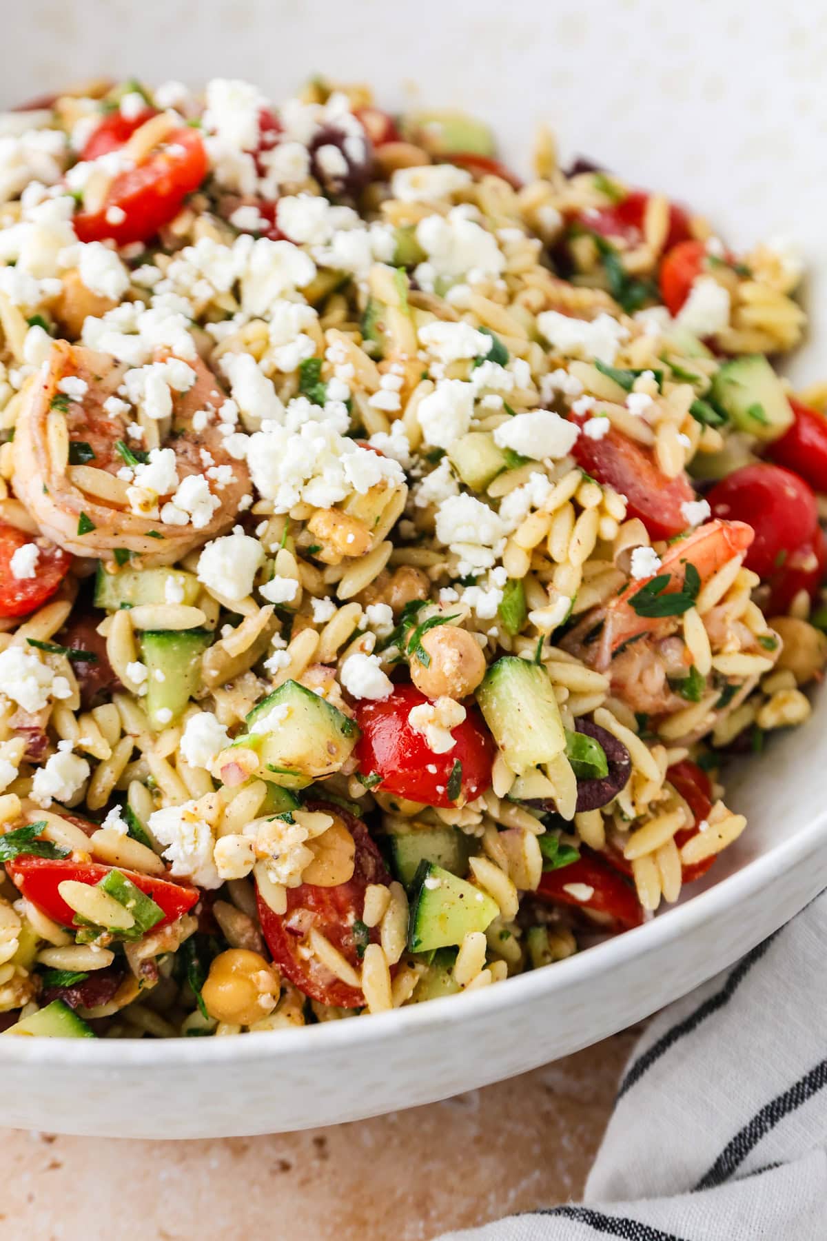 A close-up of a bowl filled with Shrimp Orzo Salad, featuring cherry tomatoes, cucumbers, olives, chickpeas, fresh herbs, and crumbled feta cheese.