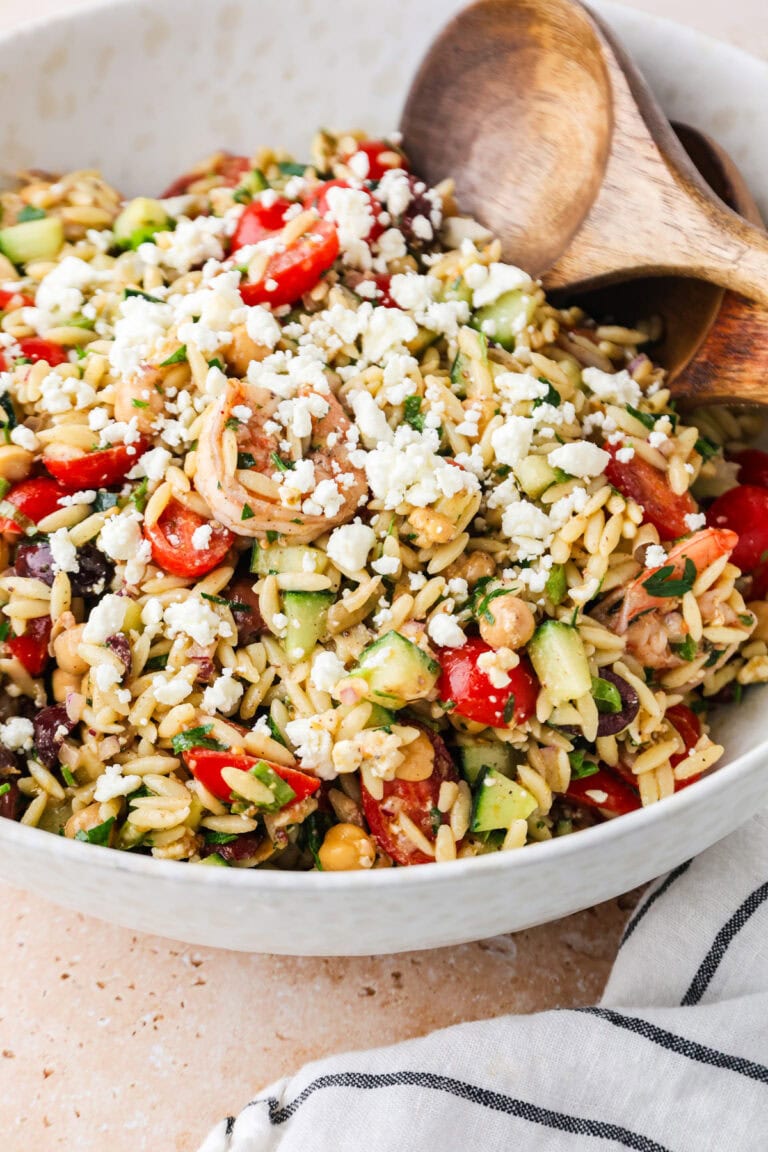 A bowl of Shrimp Orzo Salad brimming with cherry tomatoes, cucumber, chickpeas, and feta cheese, served with wooden salad tongs.