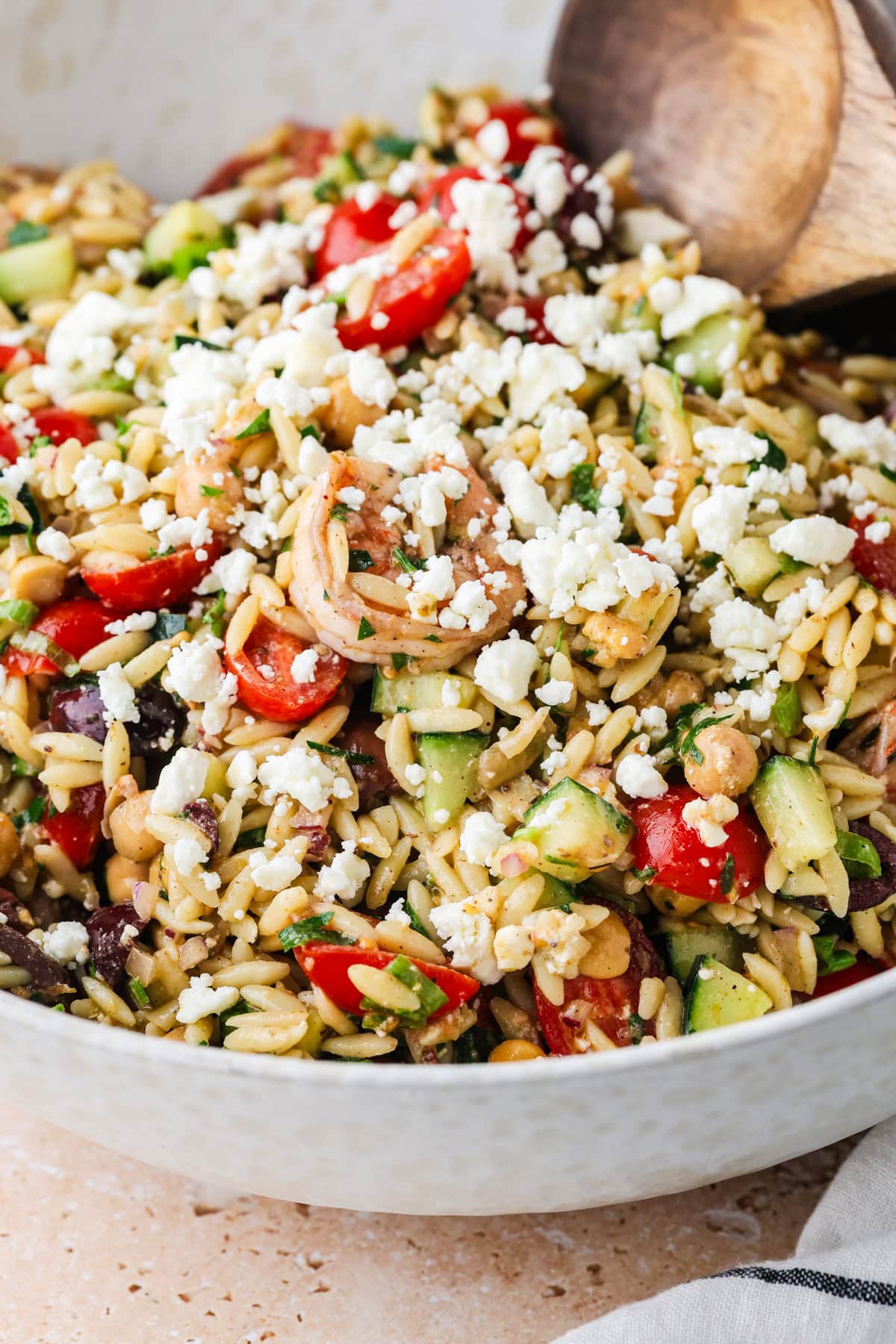 A close-up of a bowl of Shrimp Orzo Salad with chopped cucumbers, cherry tomatoes, red onions, olives, herbs, and crumbled feta cheese, with a wooden spoon resting inside the bowl.