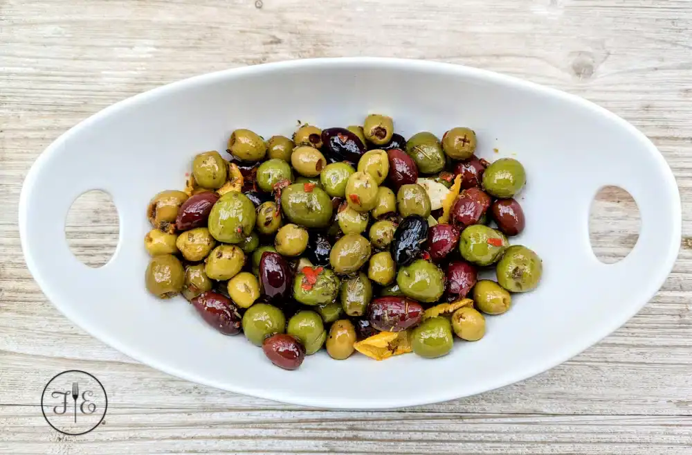 A white oval dish with handles contains a mix of green and black marinated olives, garnished with herbs and orange zest, placed on a wooden surface.