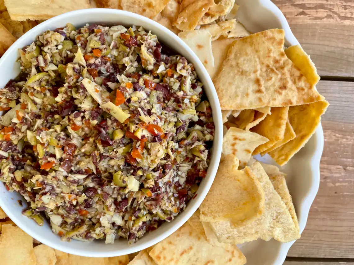 A bowl of olive tapenade with chopped vegetables is surrounded by pieces of pita chips on a white plate, sitting on a wooden surface.