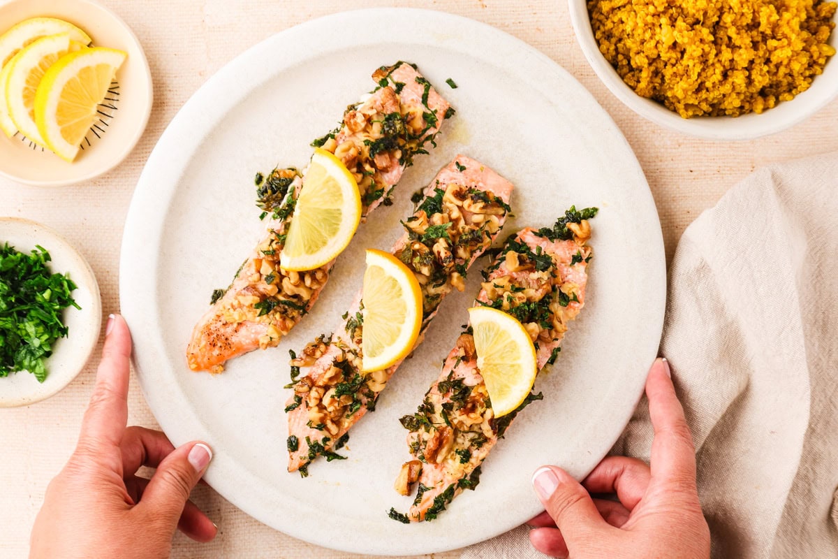 Walnut Crusted Salmon. Three salmon fillets with herbs and walnuts, each topped with a lemon wedge, served on a white plate. Sliced lemons, chopped parsley, and a bowl of quinoa are nearby. Two hands hold the plate.