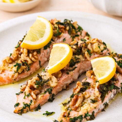 Three baked salmon fillets topped with chopped herbs, walnuts, and lemon slices are arranged on a white plate. In the background, there is a small bowl of sliced lemons and a bowl of yellow rice.
