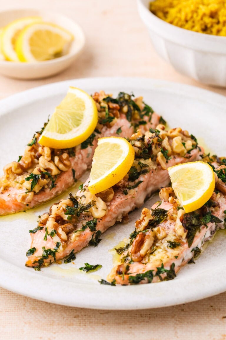 Three baked salmon fillets topped with chopped herbs, walnuts, and lemon slices are arranged on a white plate. In the background, there is a small bowl of sliced lemons and a bowl of yellow rice.