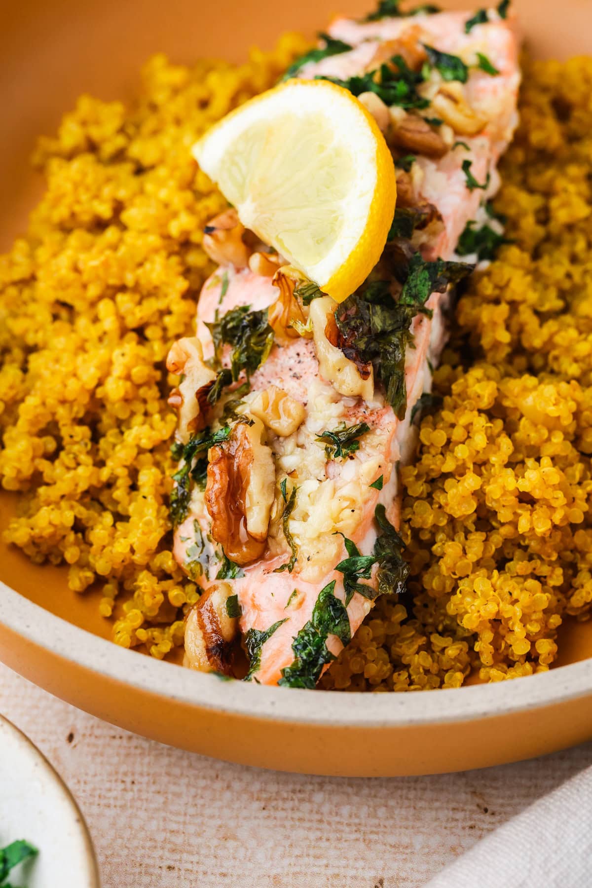 A slice of baked salmon topped with herbs, walnuts, and a lemon wedge, served on a bed of yellow quinoa in a brown bowl.