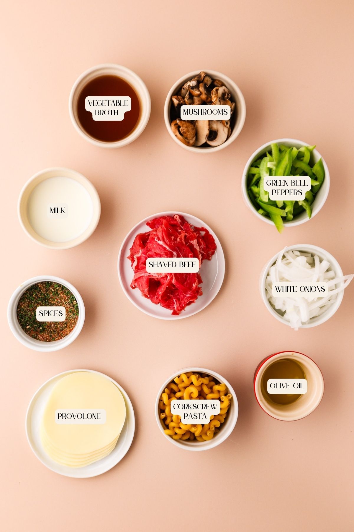 Nine bowls contain shaved beef, mushrooms, green bell peppers, white onions, spices, provolone, corkscrew pasta, milk, vegetable broth, and olive oil, all labeled and arranged on a peach background.