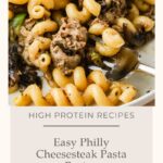 A plate of curly pasta mixed with cheesesteak-style beef, mushrooms, green peppers, and melted cheese, with a fork. Text reads: Easy Philly Cheesesteak Pasta Recipe—a delicious twist on classic Philly Cheesesteak flavors in pasta form. High Protein Recipes.