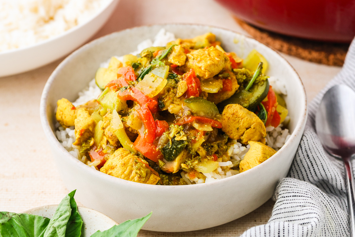 A bowl of white rice topped with colorful chicken curry containing vegetables like zucchini, tomatoes, and spinach. A spoon and napkin are beside the bowl, with a dish of rice and fresh herbs in the background.