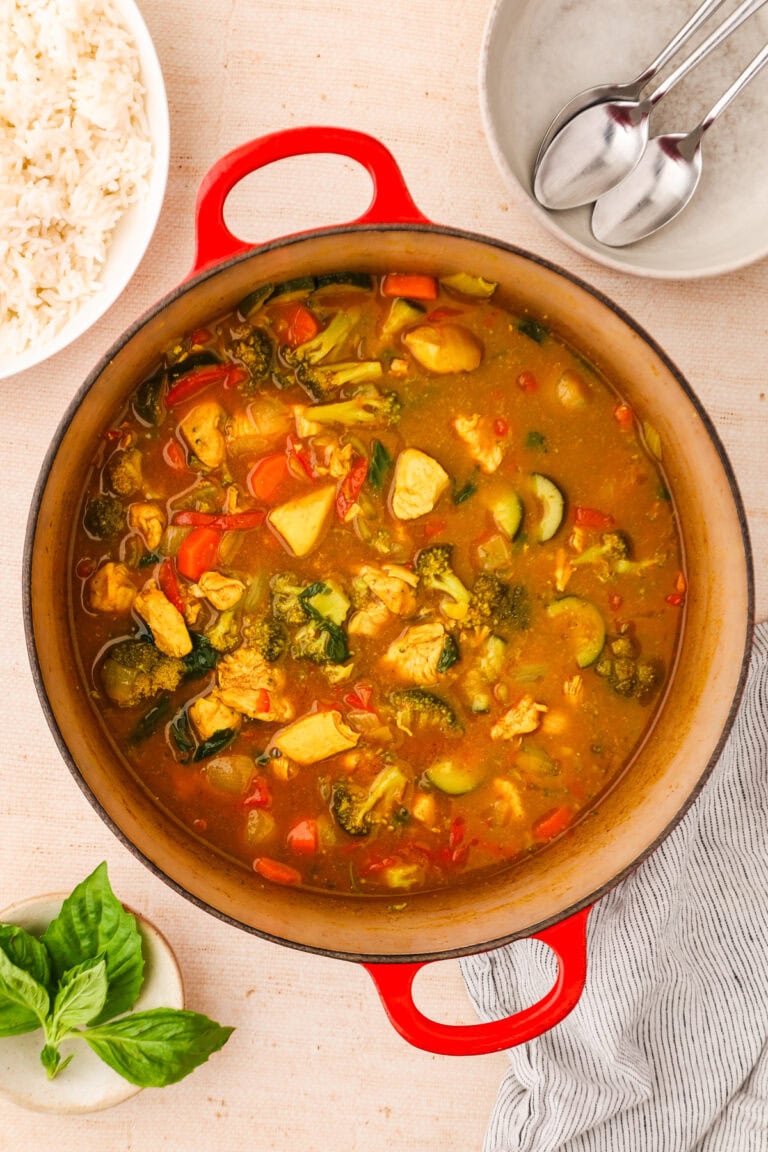 A red pot filled with chicken and vegetable curry sits on a beige surface, surrounded by a bowl of rice, a plate with spoons, fresh basil, and a striped napkin.